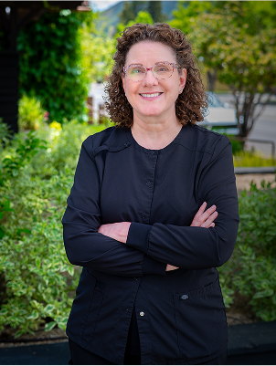A person with curly hair and glasses smiles confidently with arms crossed, standing outside. Lush greenery and a street are visible in the background.