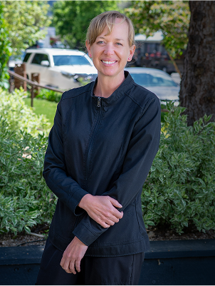 A person in a black jacket smiles warmly while standing in front of green foliage. A white vehicle is visible in the blurred background, suggesting an outdoor setting.