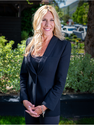 Blonde woman in a black blazer smiles confidently outdoors, with greenery and parked cars in the background. Bright, sunny day.
