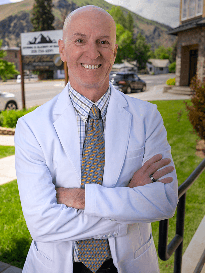 Dr. Roth in a white coat stands outside, arms crossed, smiling confidently. Background includes greenery, a sign, buildings, and mountains in the distance.