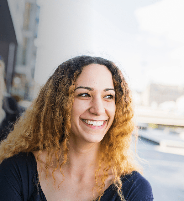 A woman with curly, golden-brown hair smiles brightly outdoors. She wears a navy top, projecting joy and warmth under a clear sky, with blurred urban surroundings.