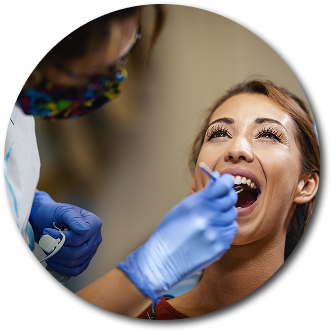 A woman smiles with mouth open during a dental check-up. A dentist, in blue gloves, inspects her teeth. The atmosphere is professional and relaxed.