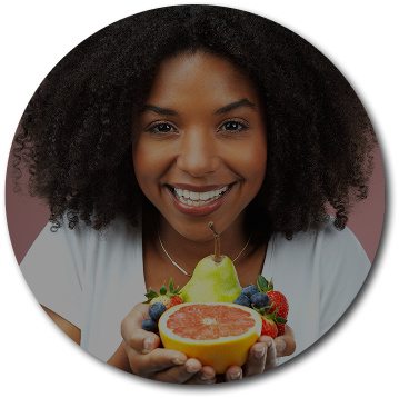 Smiling woman with curly hair holds fresh fruit, including a pear, grapefruit, strawberries, and blueberries, against a pink background.