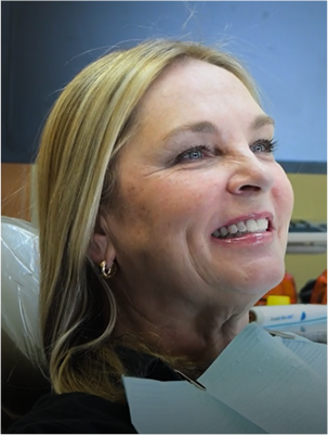 Smiling woman with blonde hair sits in a dentist's chair, wearing a dental bib. Background shows dental tools, creating a cheerful, relaxed atmosphere.