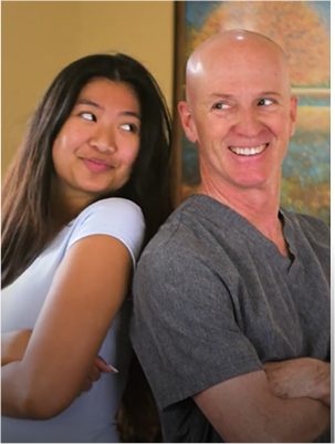 A client and Dr Jeff stand back-to-back, smiling at each other in a warm, brightly-lit room with a painting in the background.