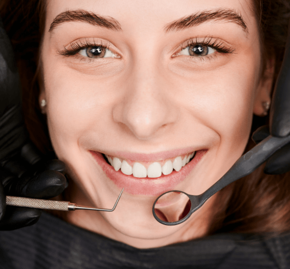 A smiling woman is at the dentist, with her teeth being examined. The dentist's gloved hands hold a mirror and a dental tool. The mood is cheerful.