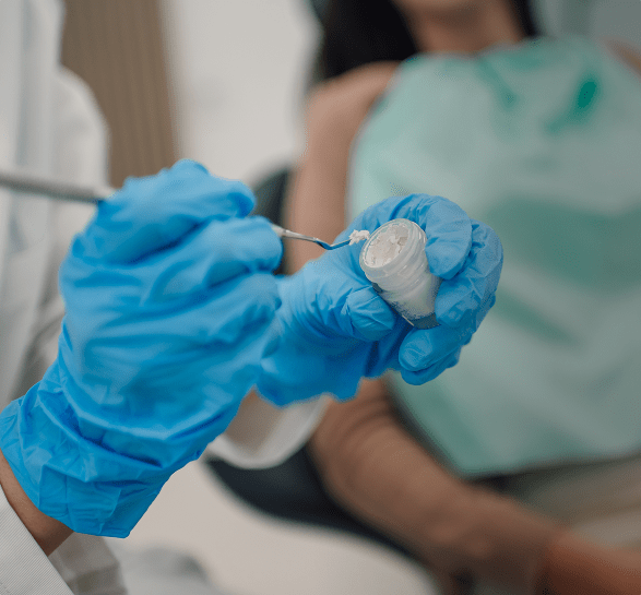 A dentist wearing blue gloves prepares dental material with a tool. A patient, blurred in the background, wears a green bib, evoking a clinical setting.