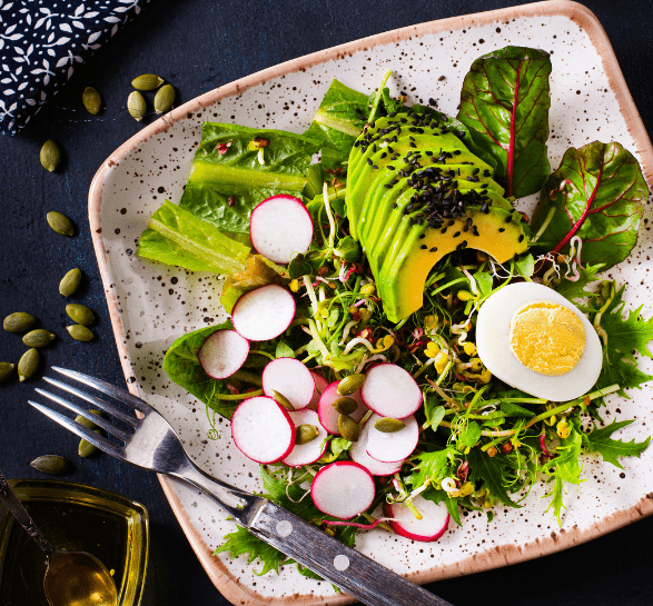 A fresh salad on a speckled plate with avocado slices, radish, greens, and a halved boiled egg. Topped with seeds, exuding a vibrant, healthy vibe.
